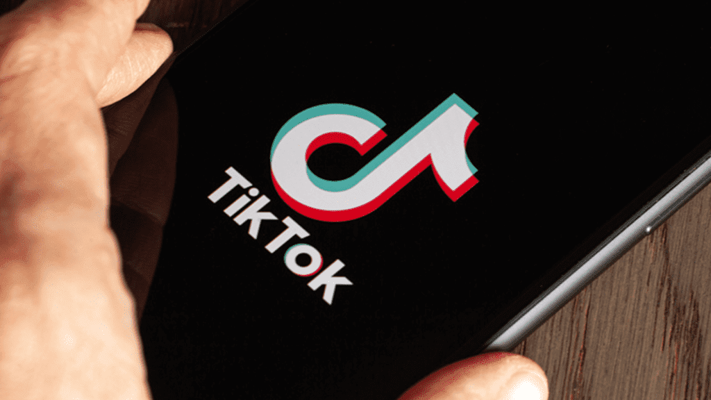 Aparelho celular encima de uma mesa de madeira com a logo do TikTok na tela e uma mão próxima ao aparelho.