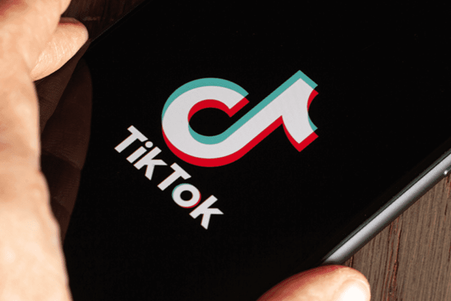 Aparelho celular encima de uma mesa de madeira com a logo do TikTok na tela e uma mão próxima ao aparelho.